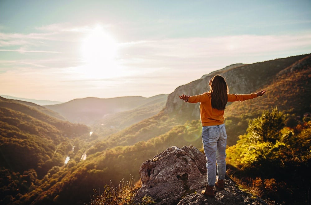 Person standing on a rocky overlook with arms outstretched, facing a sunlit mountain landscape with autumn-colored trees.