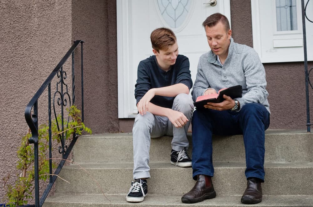 Adult and teenage boy sitting on front steps, looking at a Bible together and talking.