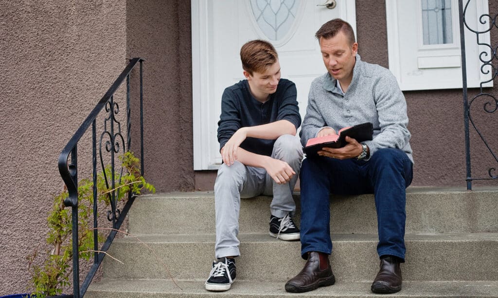 Adult and teenage boy sitting on front steps, looking at a Bible together and talking.