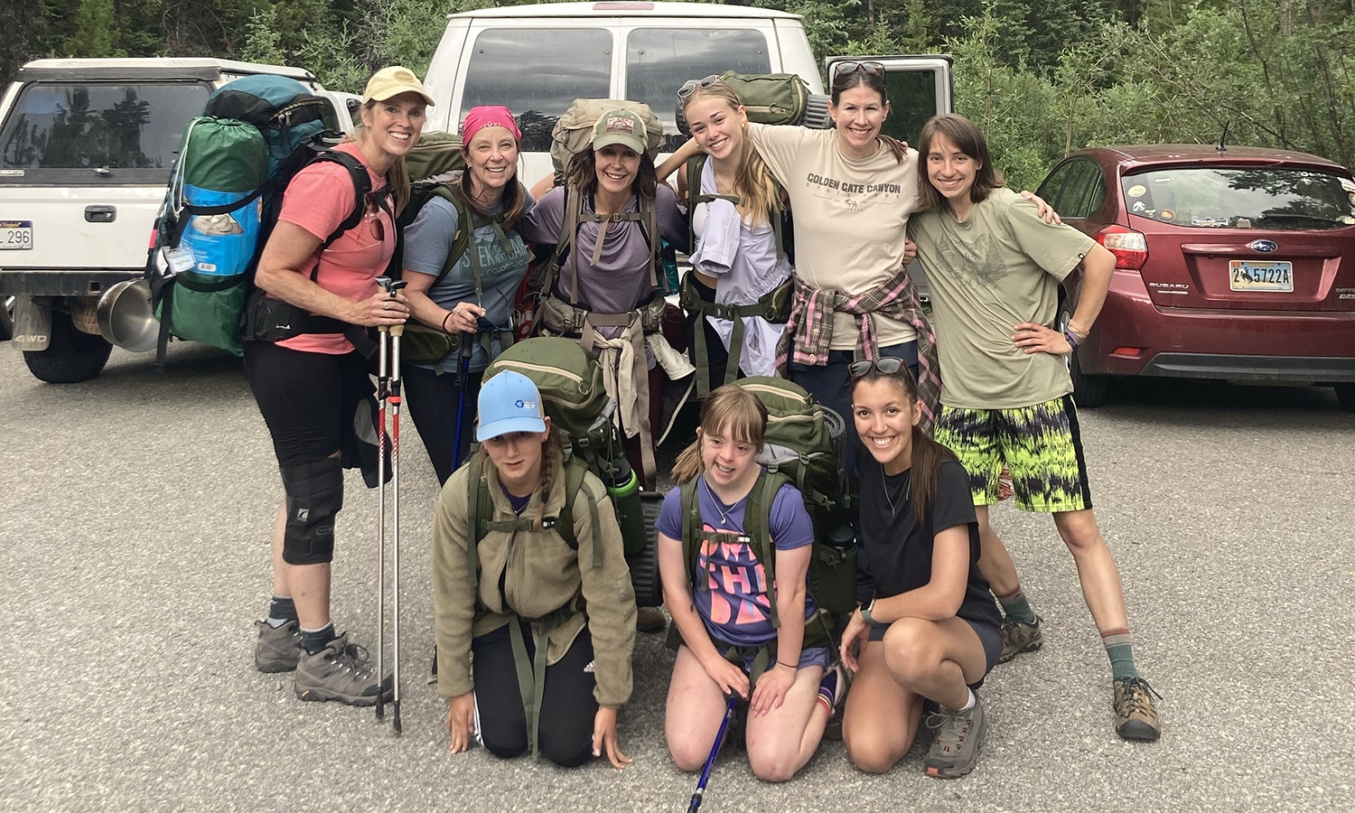 A group of nine hikers pose together in a forest parking area, smiling in front of a white van. They are wearing backpacks and outdoor gear, with some holding trekking poles.